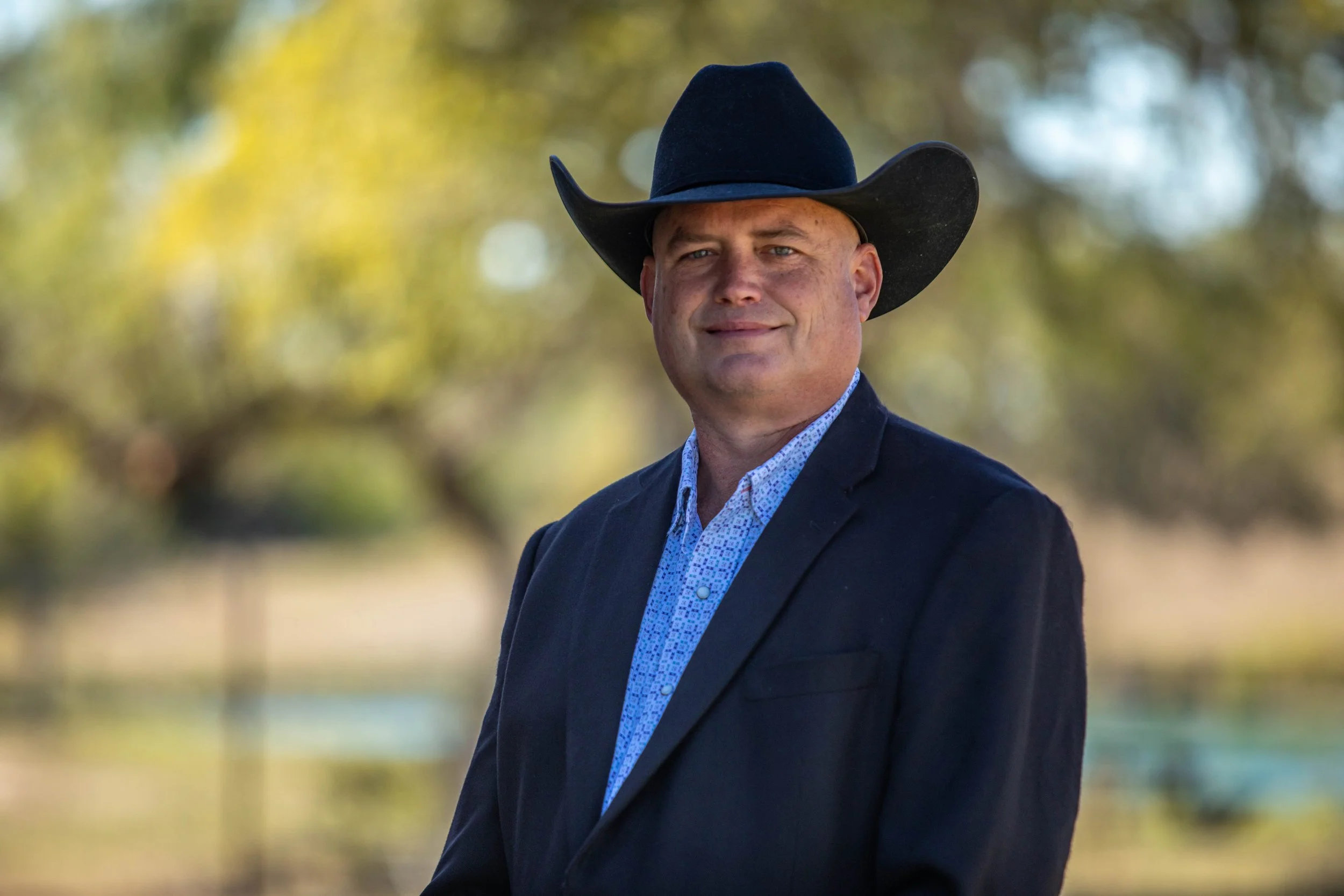 A man wearing a black cowboy hat, dark blazer, and a light patterned shirt outdoors with trees and blurred background.