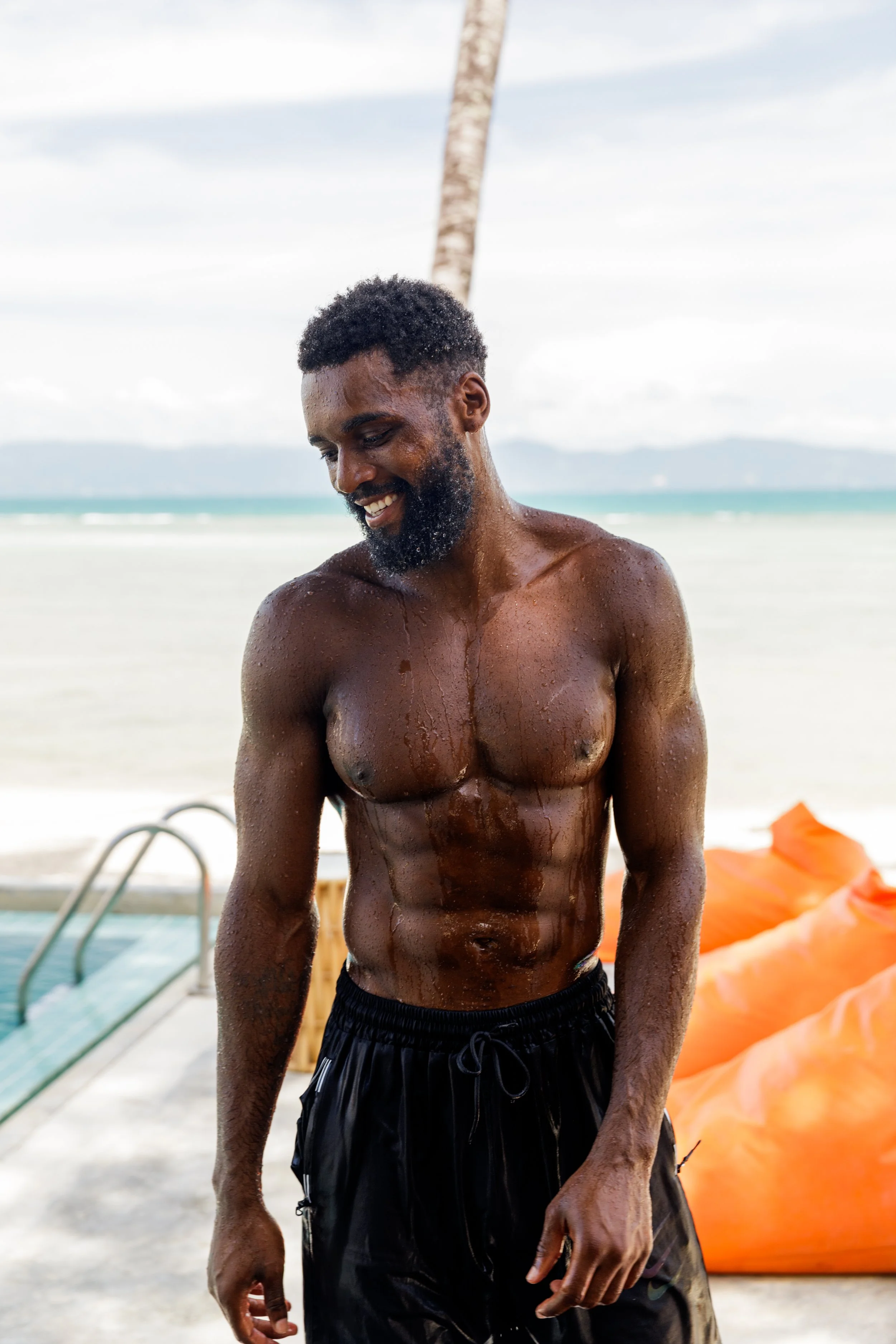 A smiling, shirtless man with wet skin and curly black hair standing on a beach with lounge chairs and a palm tree in the background.