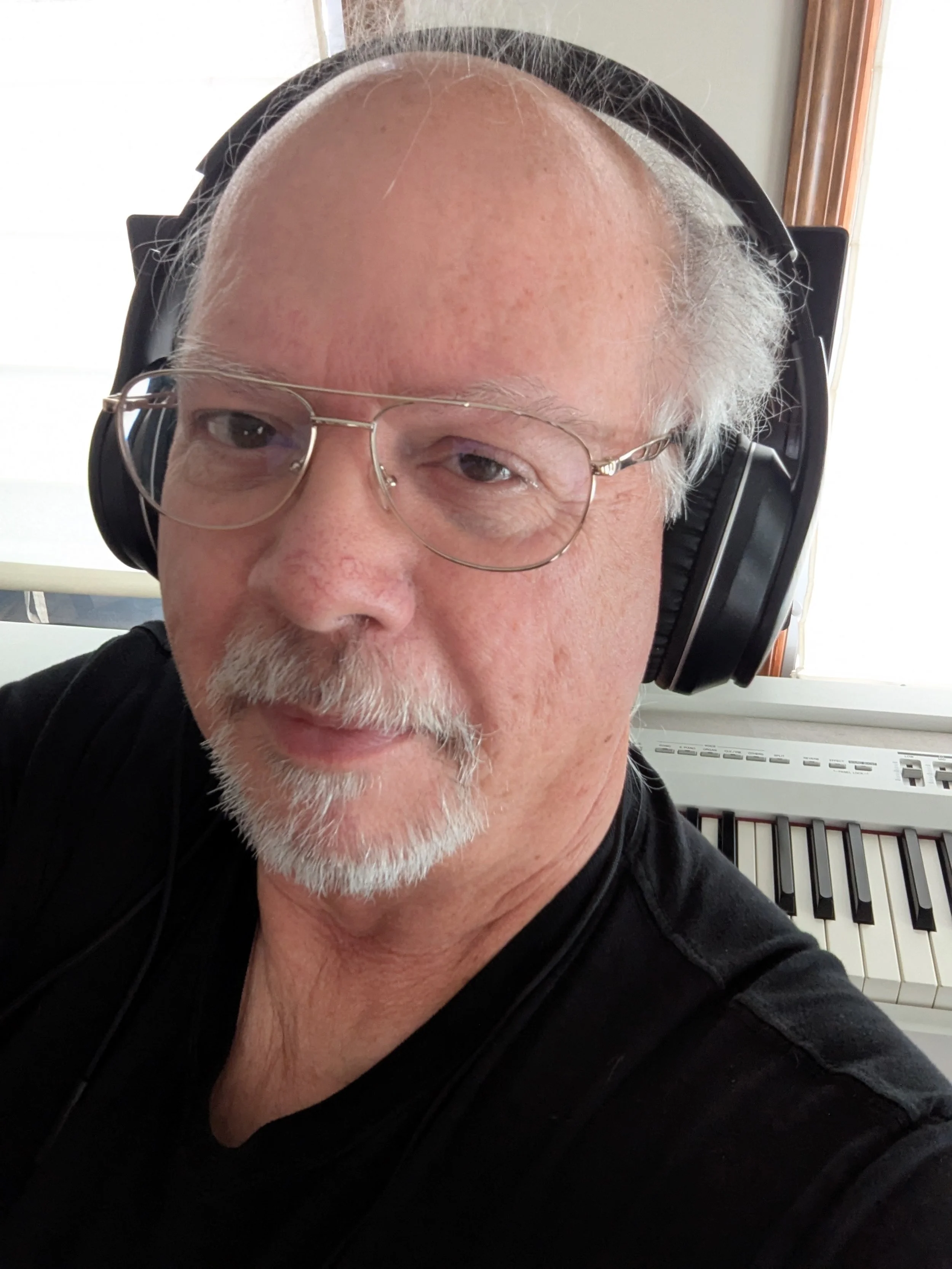 Close-up of a mature man with glasses and white facial hair, wearing large headphones, sitting in front of a keyboard piano.