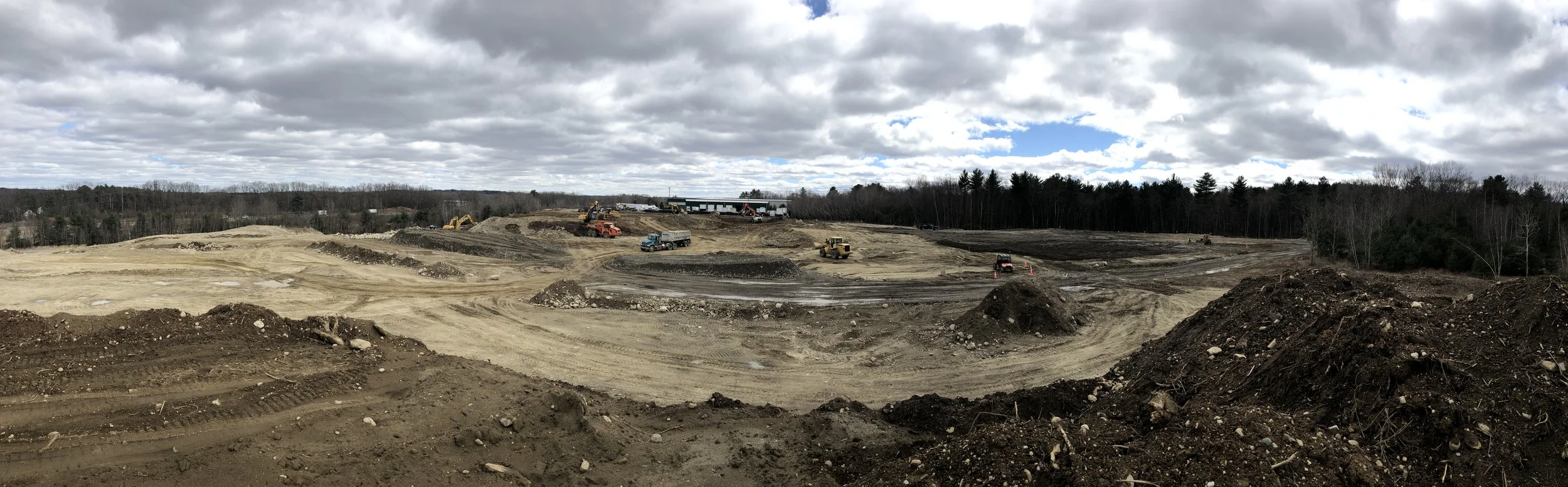 Construction site with dirt mounds, construction vehicles, and cloudy sky.