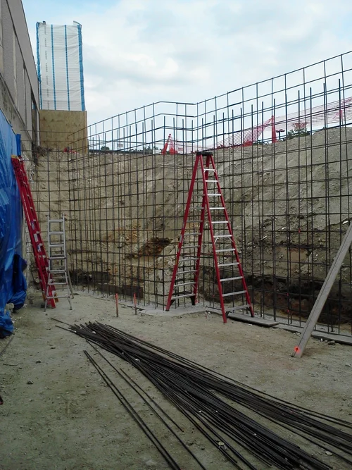 Construction site with steel rebar installed and several ladders, with a deep excavation and construction materials on the ground.