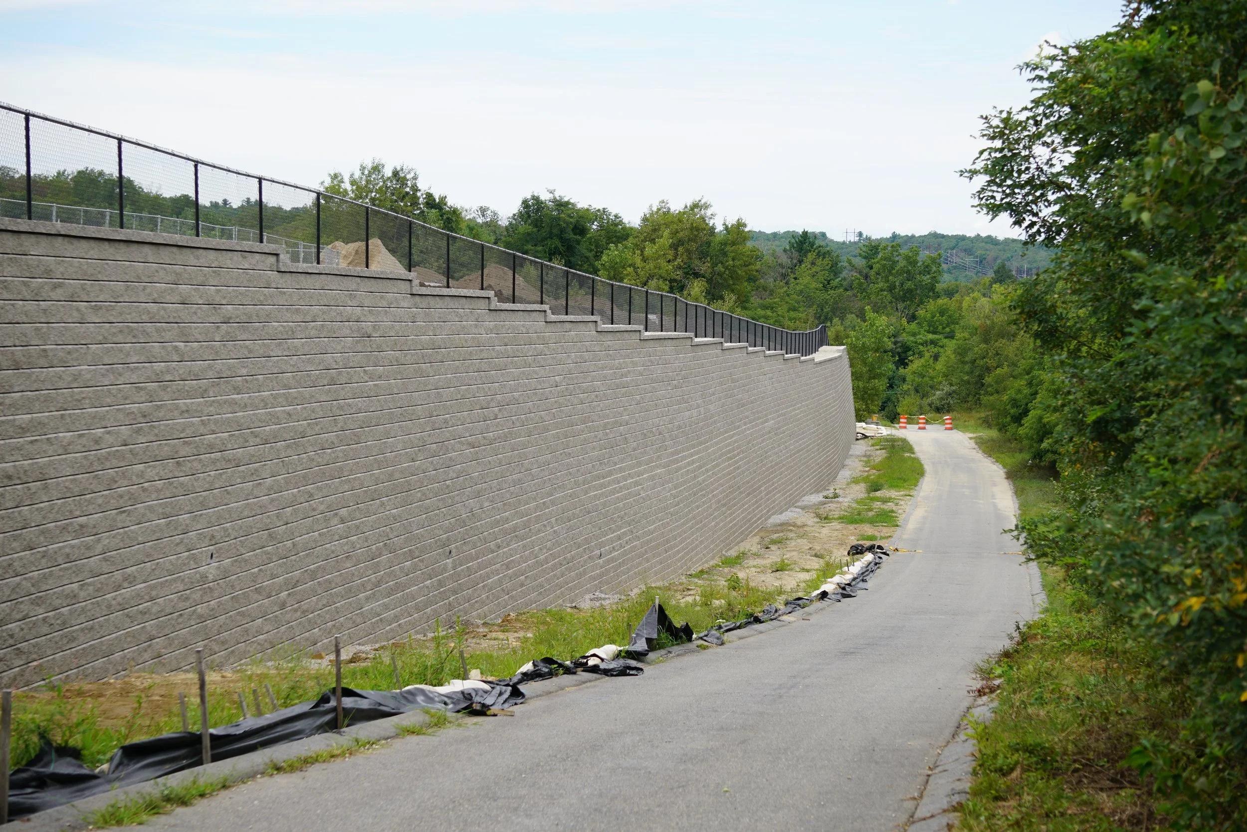 Construction of a large concrete retaining wall alongside a narrow road in a green, hilly area with trees. Construction materials and black silt fences are visible along the base of the wall.