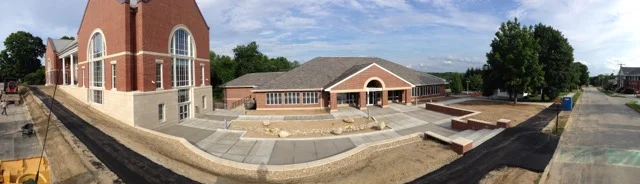 Panoramic view of a church under construction, with a brick tower, a main building with a brown roof, concrete pathways, and landscaping in progress.