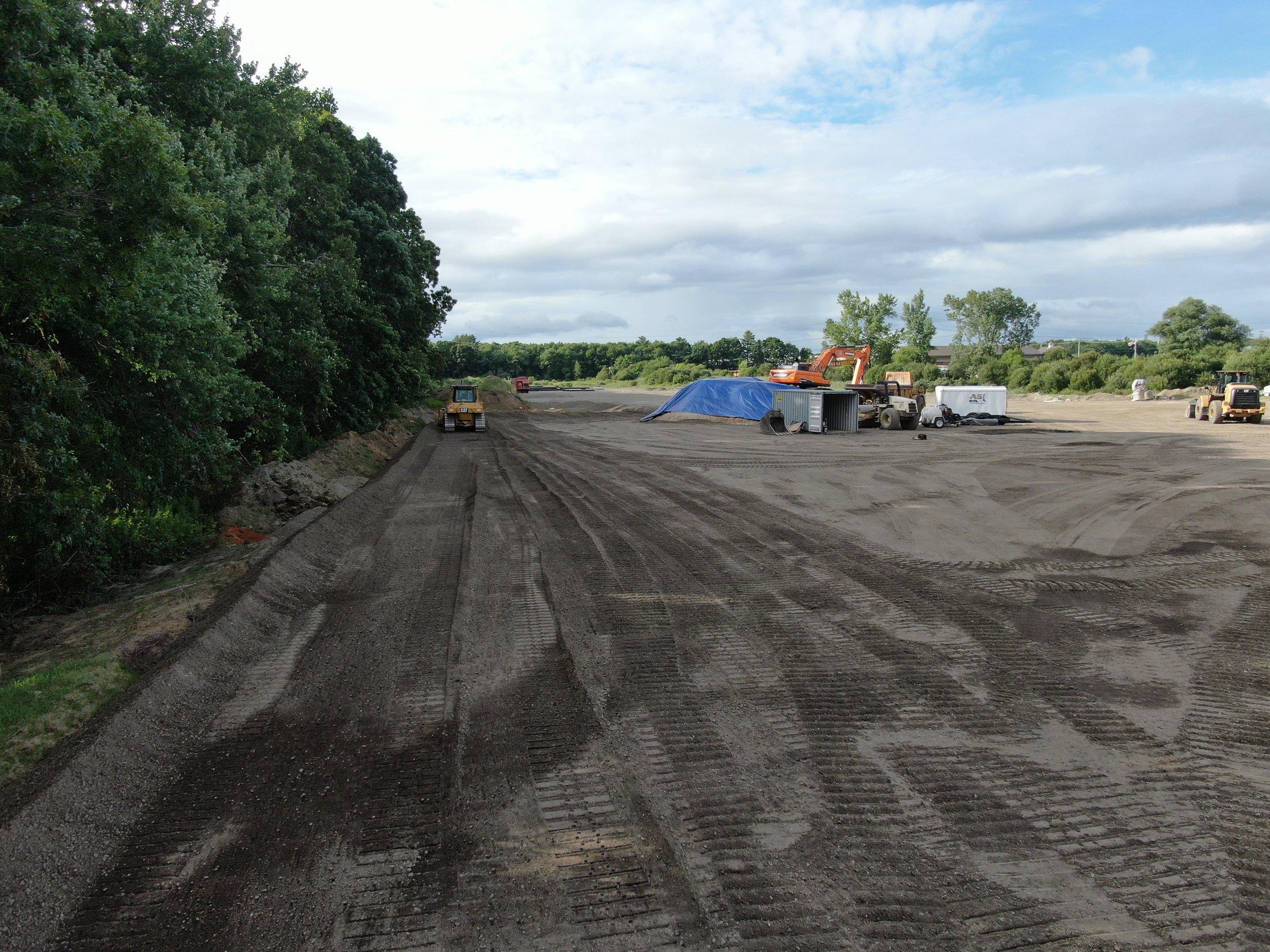 Construction site with heavy machinery and equipment on a partially finished road, surrounded by greenery and open sky.
