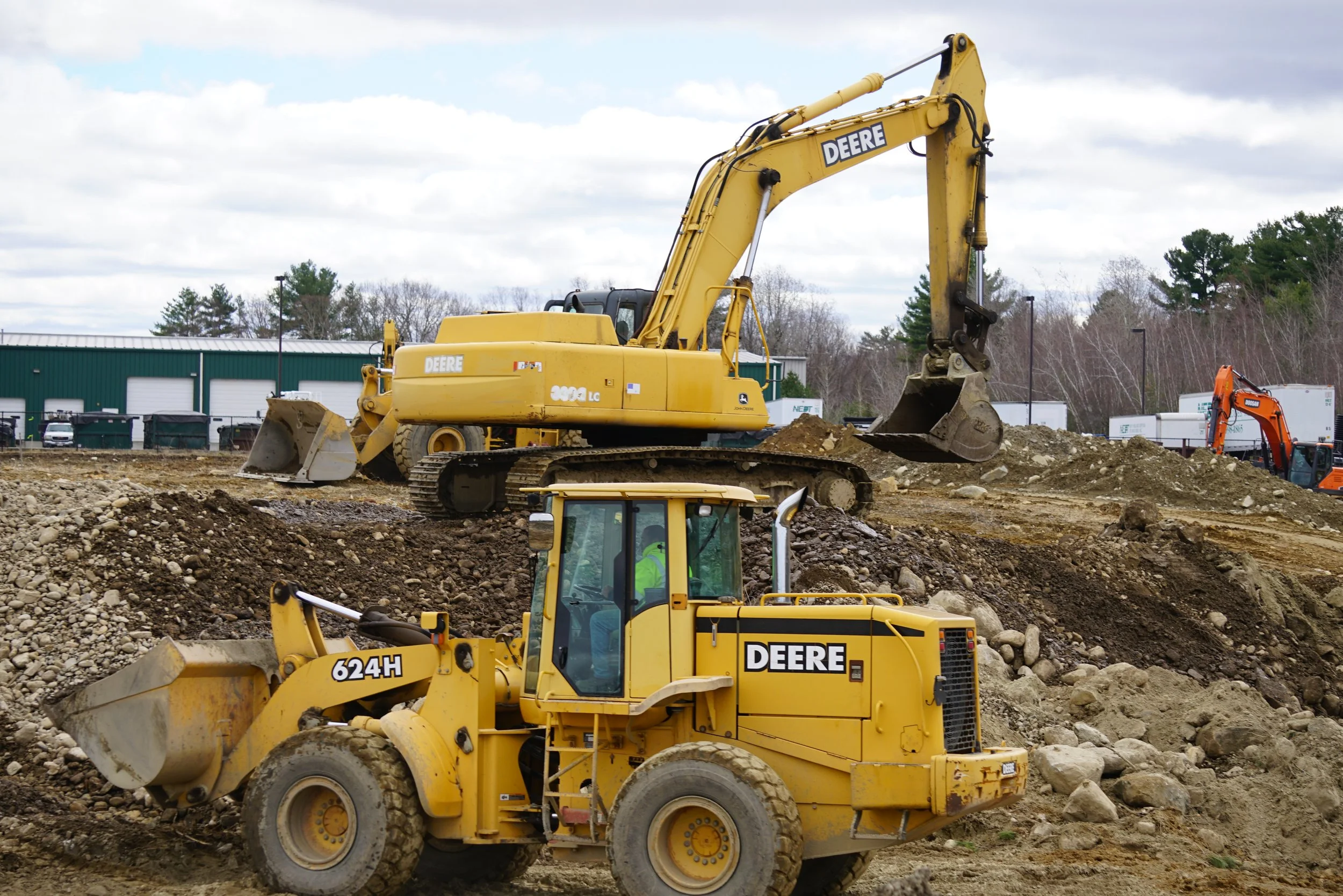 Construction site with multiple yellow and orange John Deere heavy machinery including excavators and loaders working on dirt and rocks under a cloudy sky, with a green building and trees in the background.