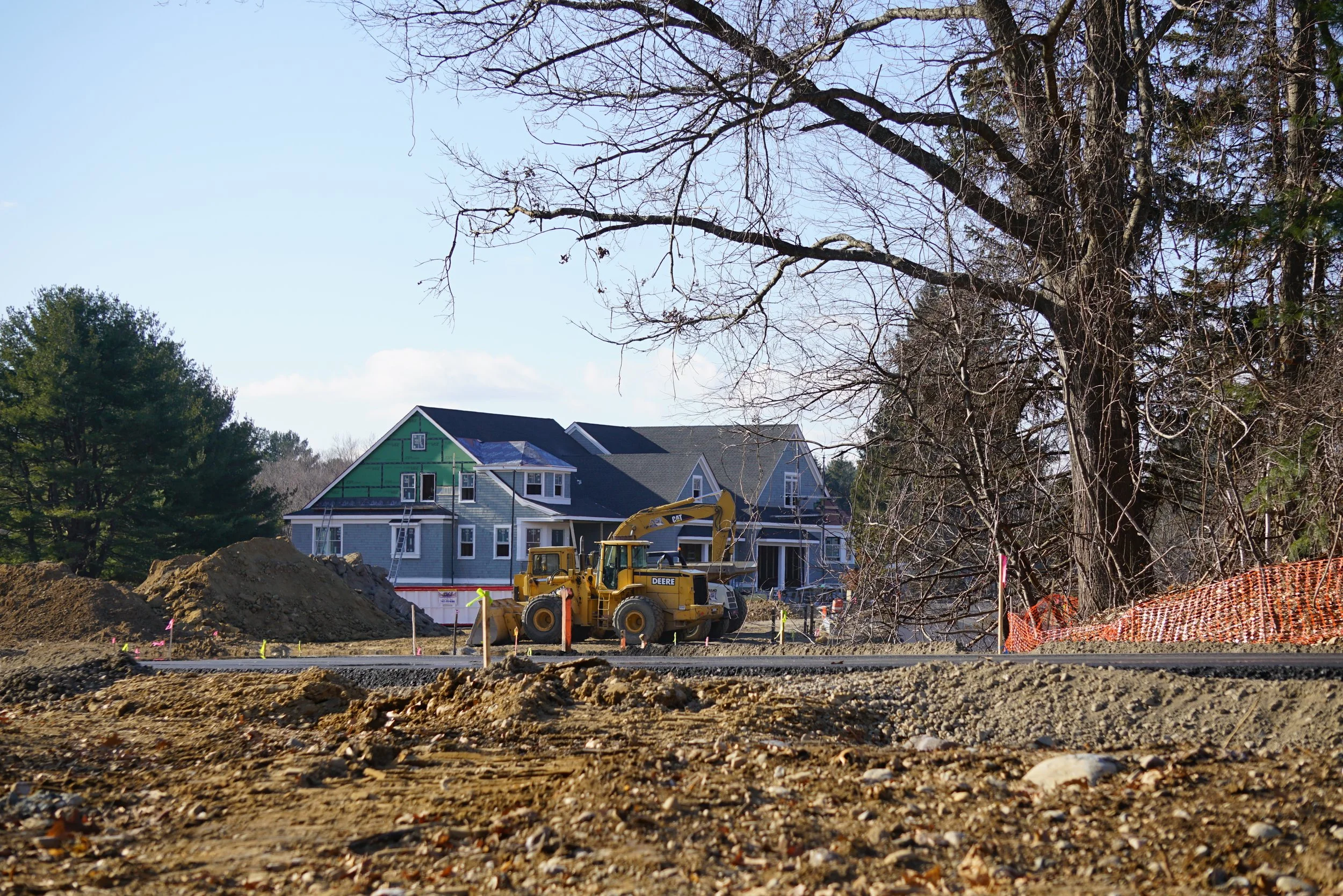 Construction site with a yellow excavator and piles of dirt, residential houses in the background, and trees with mostly bare branches.