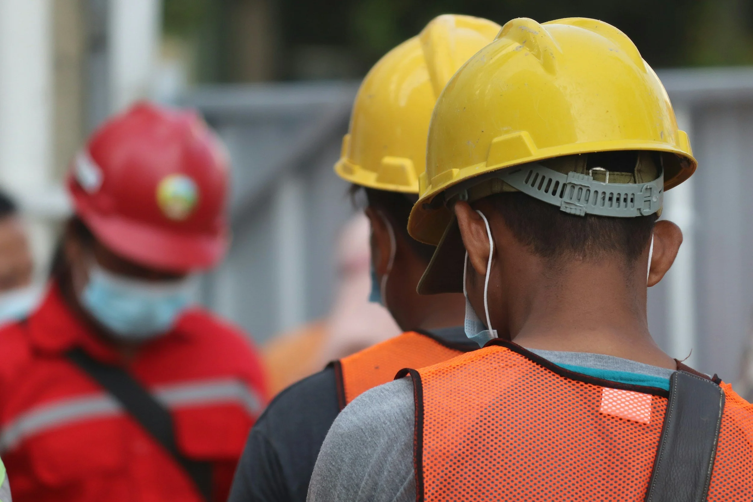 People wearing yellow and red safety helmets and face masks, with some in orange and gray clothing, likely construction workers, gathered outdoors.