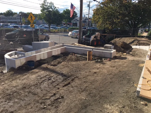 Construction site with a partially built concrete outline, construction equipment, and an American flag in the background.