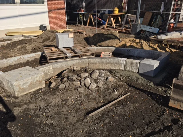 Construction site with large stone blocks forming a curved border, dirt and rocks on the ground, construction tools, and materials scattered around.