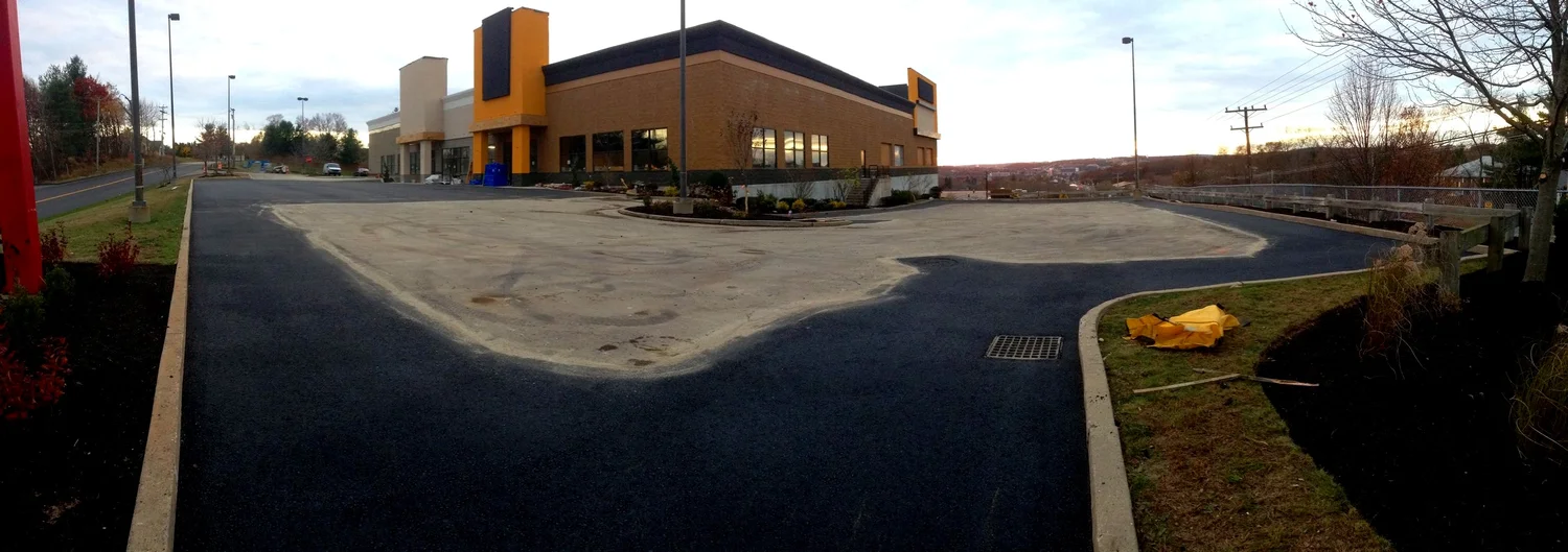 Newly paved asphalt parking lot and the exterior of a fast food restaurant during dusk with trees and a cloudy sky in the background.