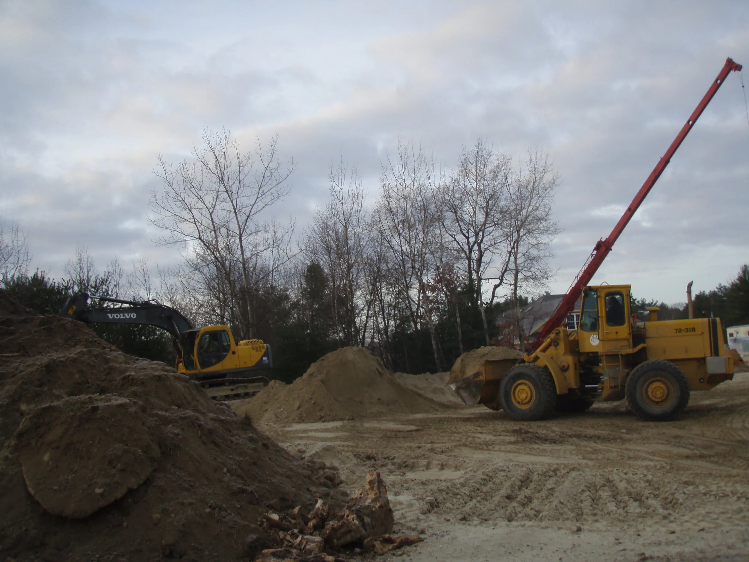 Construction site with excavator and dirt piles, with a cloudy sky and leafless trees in the background.