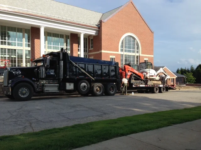 A large black tow truck is parked in front of a brick building with large windows. The truck is carrying a flatbed trailer with several white cars and construction equipment on it. A person is standing near the trailer.
