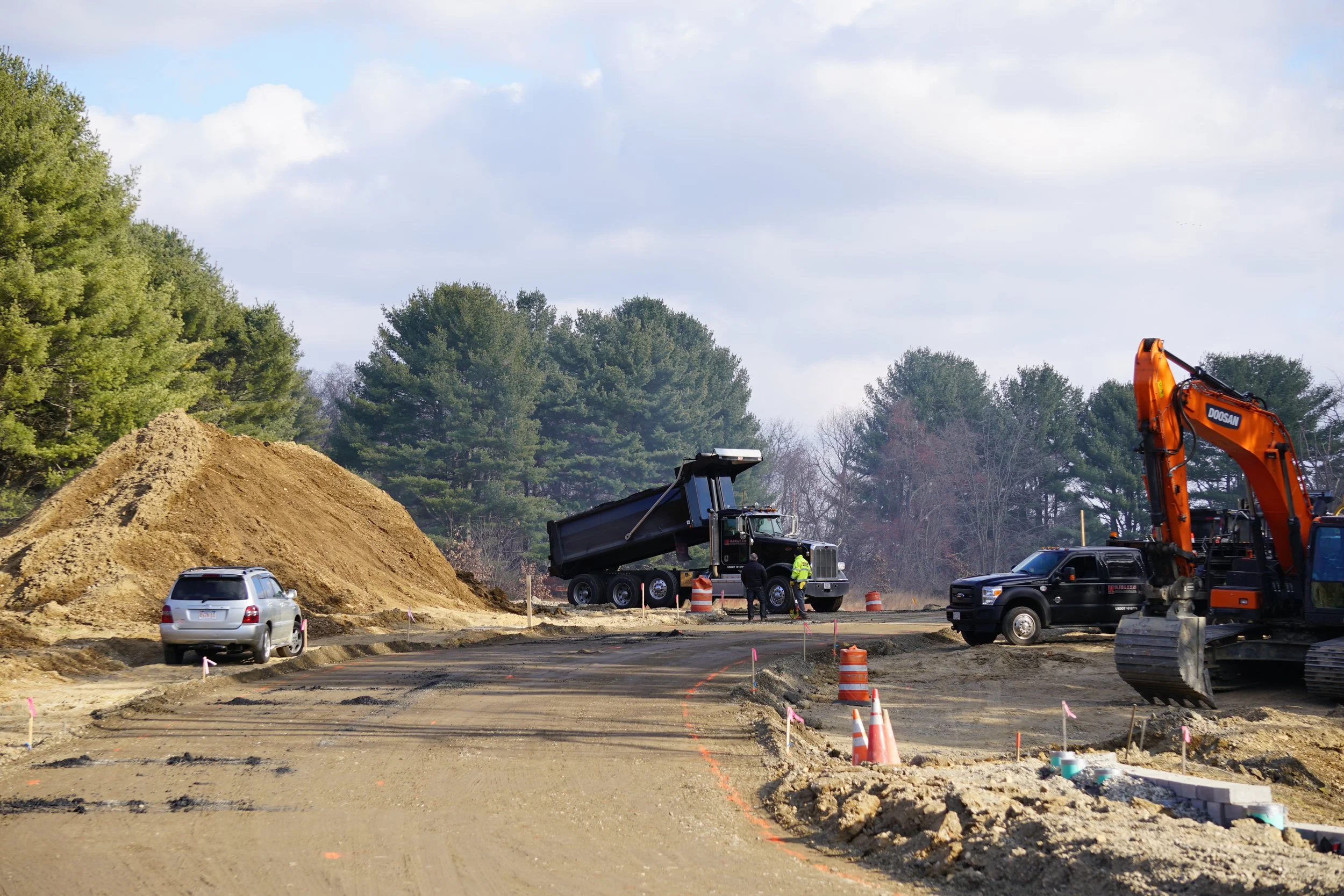 Construction site with heavy machinery, dirt piles, and workers. An excavator and dump truck are actively working, with orange safety cones and markers on the ground, and trees and a cloudy sky in the background.