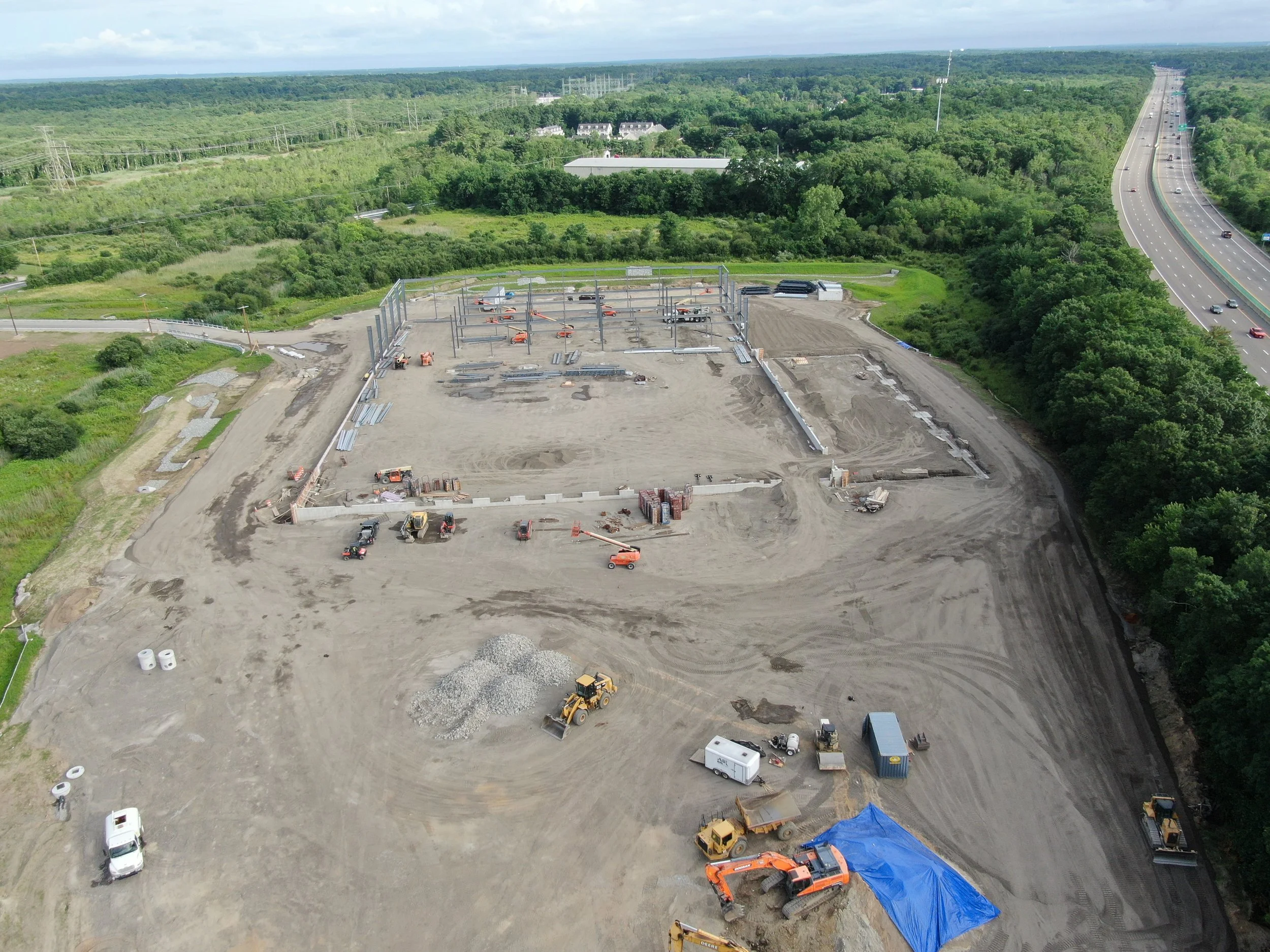 Aerial view of a construction site with building framework, construction vehicles, and materials, surrounded by greenery and a highway with moving cars.