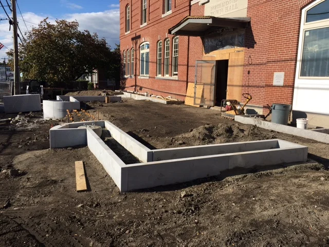 Construction site with fresh concrete footings for landscaping or building extension outside a brick building.