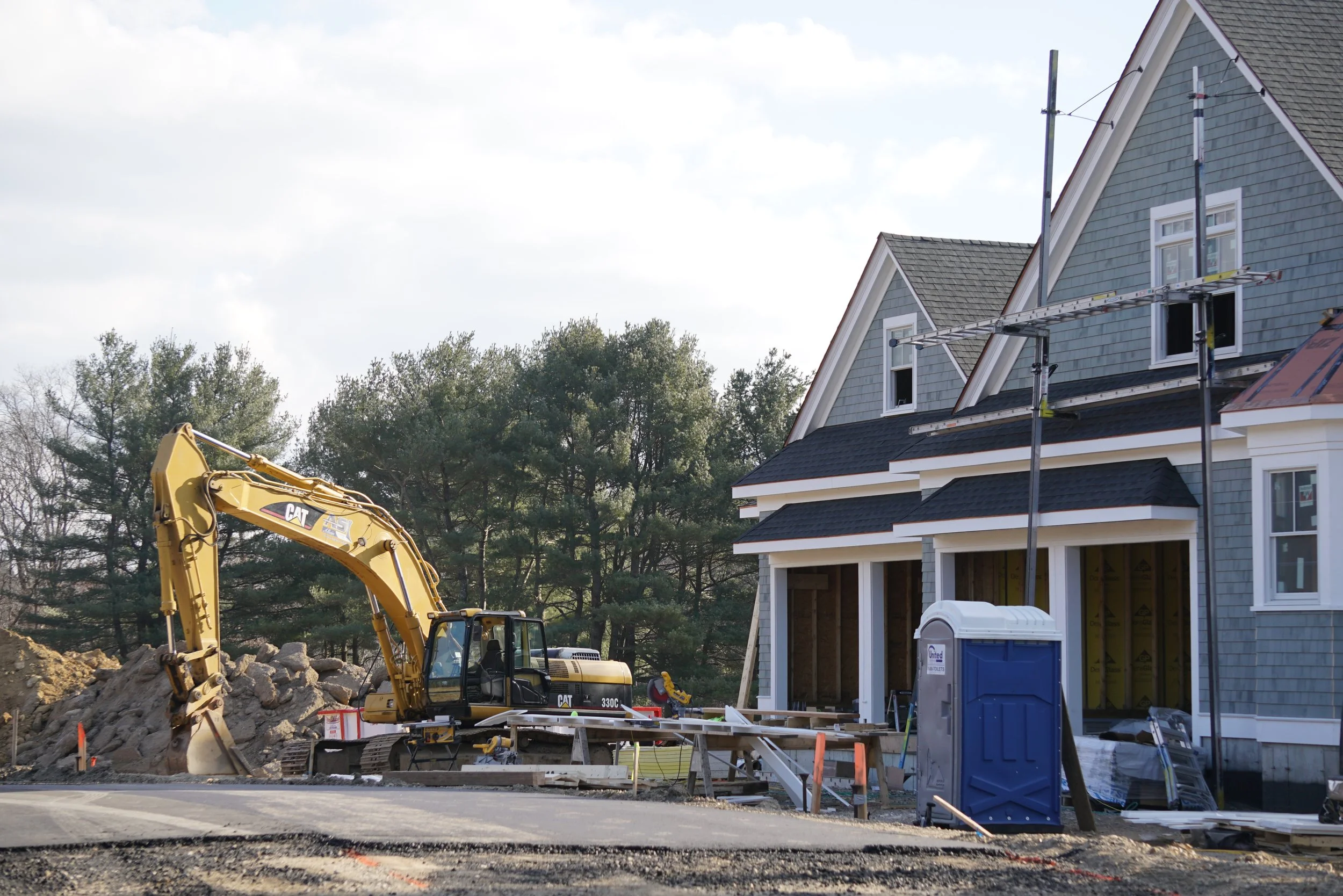 A construction site with a yellow excavator, a partially built house with scaffolding, USA Toilets portable toilet, and a pile of dirt and rocks, with trees in the background.
