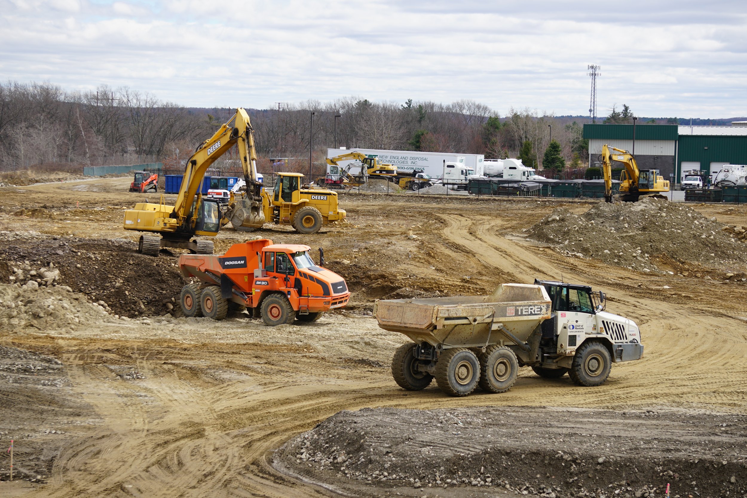 Construction site with heavy machinery including bulldozers, excavators, and a dump truck, with a building and trucks in the background and trees in the distance under a cloudy sky.