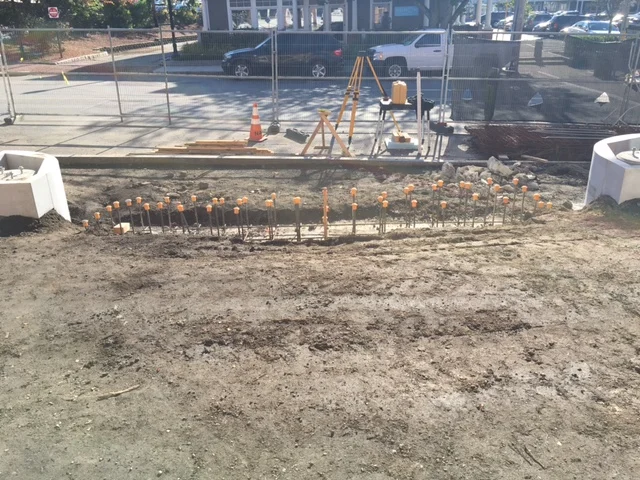 Construction site with rebar and orange caps for future concrete work, orange cones, and construction fencing in a parking lot.
