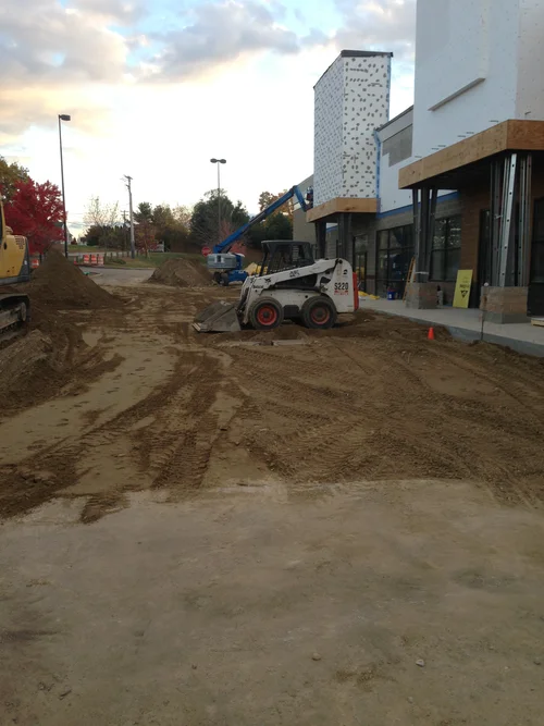 Construction site with dirt and heavy machinery near a building with a white and brown facade