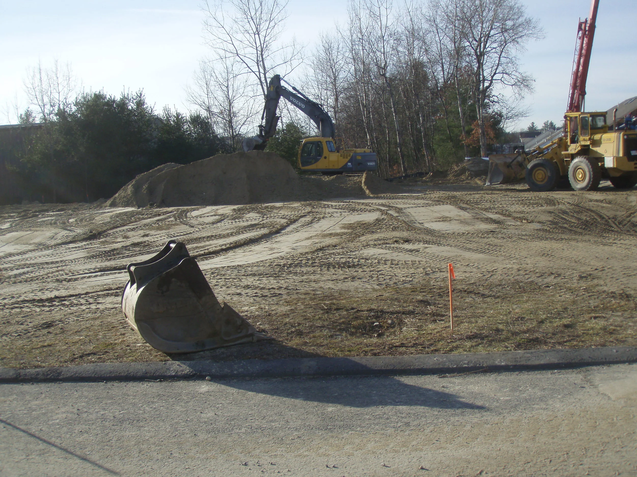 Construction site with a yellow excavator loading dirt onto a pile, a bulldozer nearby, and tire tracks in the dirt, with leafless trees in the background.