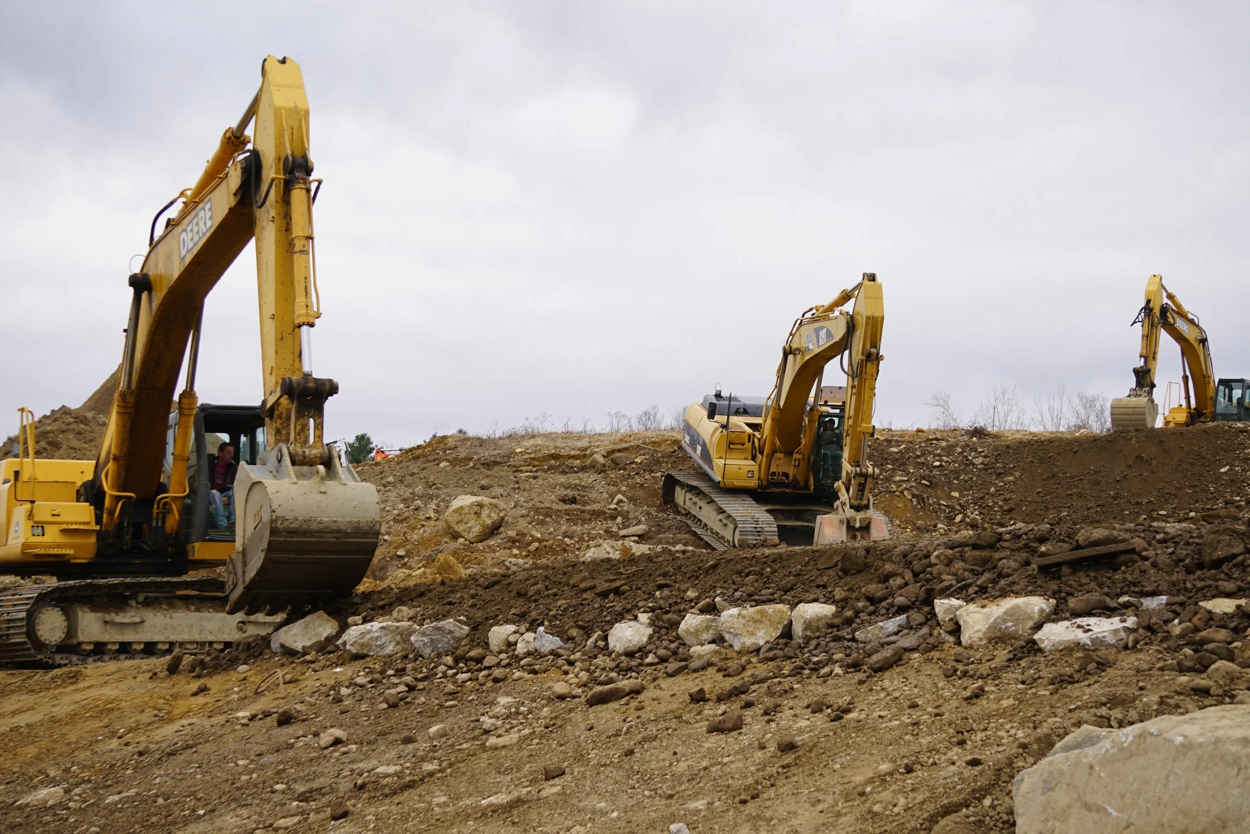 Multiple yellow construction excavators working on a rocky and dirt-covered construction site under an overcast sky.