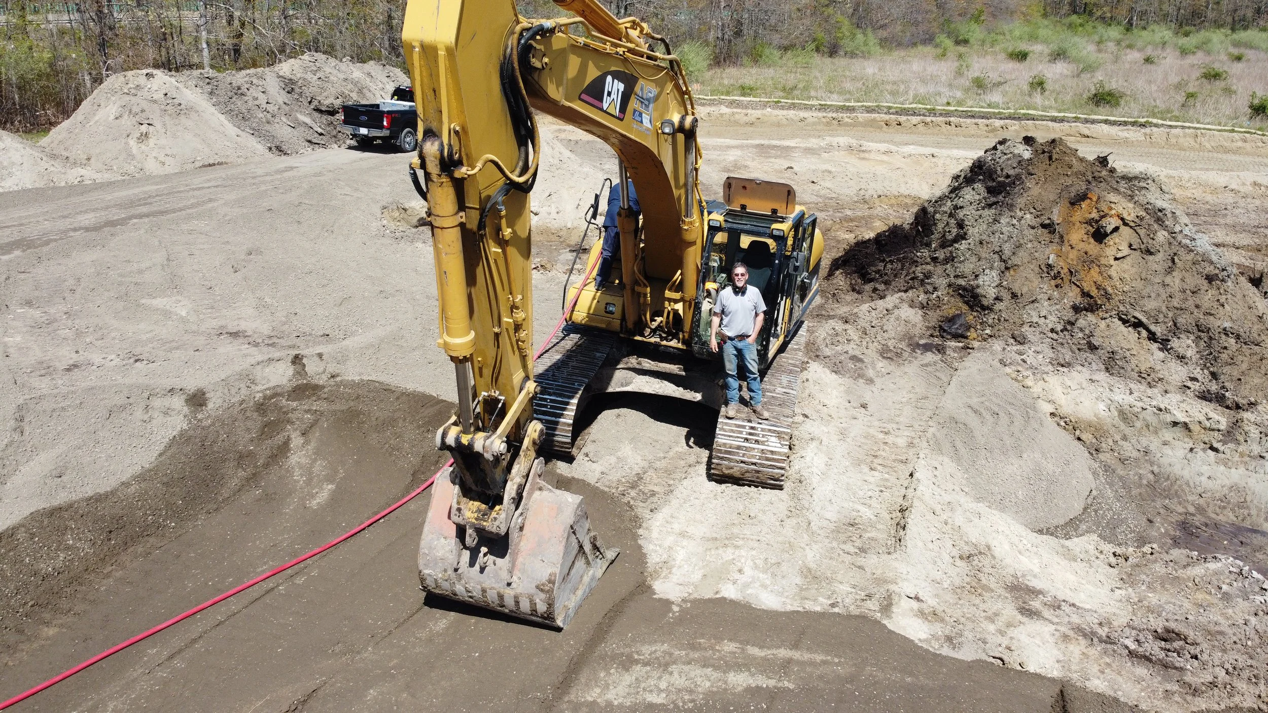 A man standing on a construction site next to a large yellow excavator, with a pile of dirt and a black pickup truck in the background.
