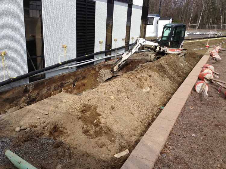 Construction site with a small excavator digging a trench alongside a building, with construction barriers on the sidewalk.