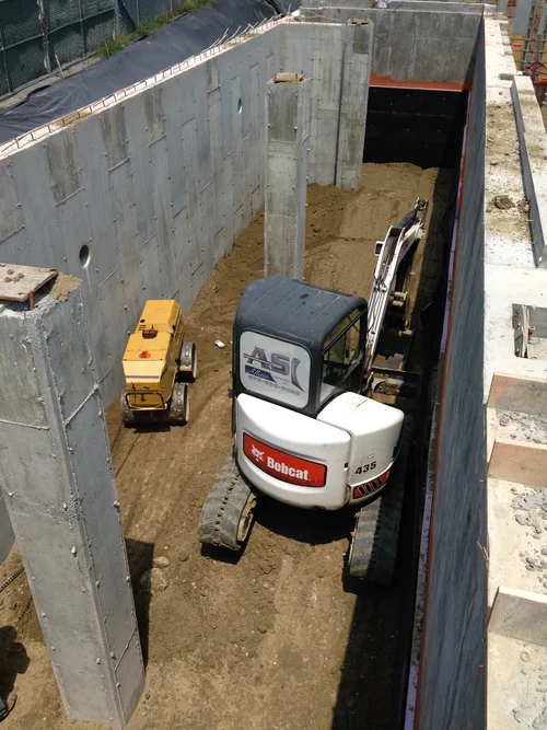 Construction site with Bobcat mini excavator and small yellow compactor inside deep concrete foundation walls.