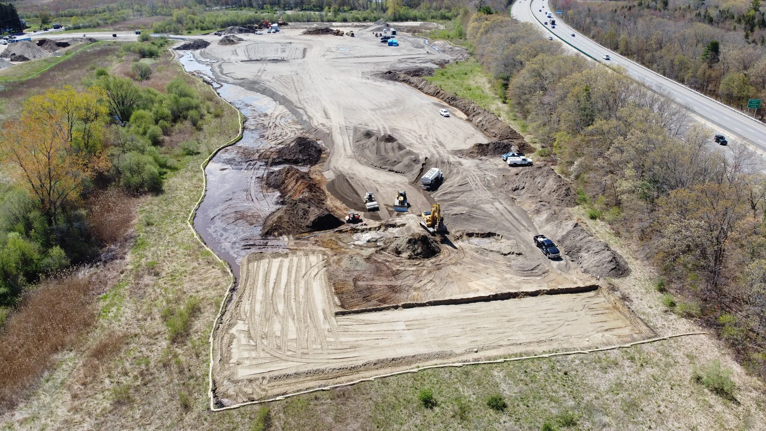 Construction site along a river with earth-moving equipment and vehicles, next to a highway with traffic, surrounded by trees and greenery.