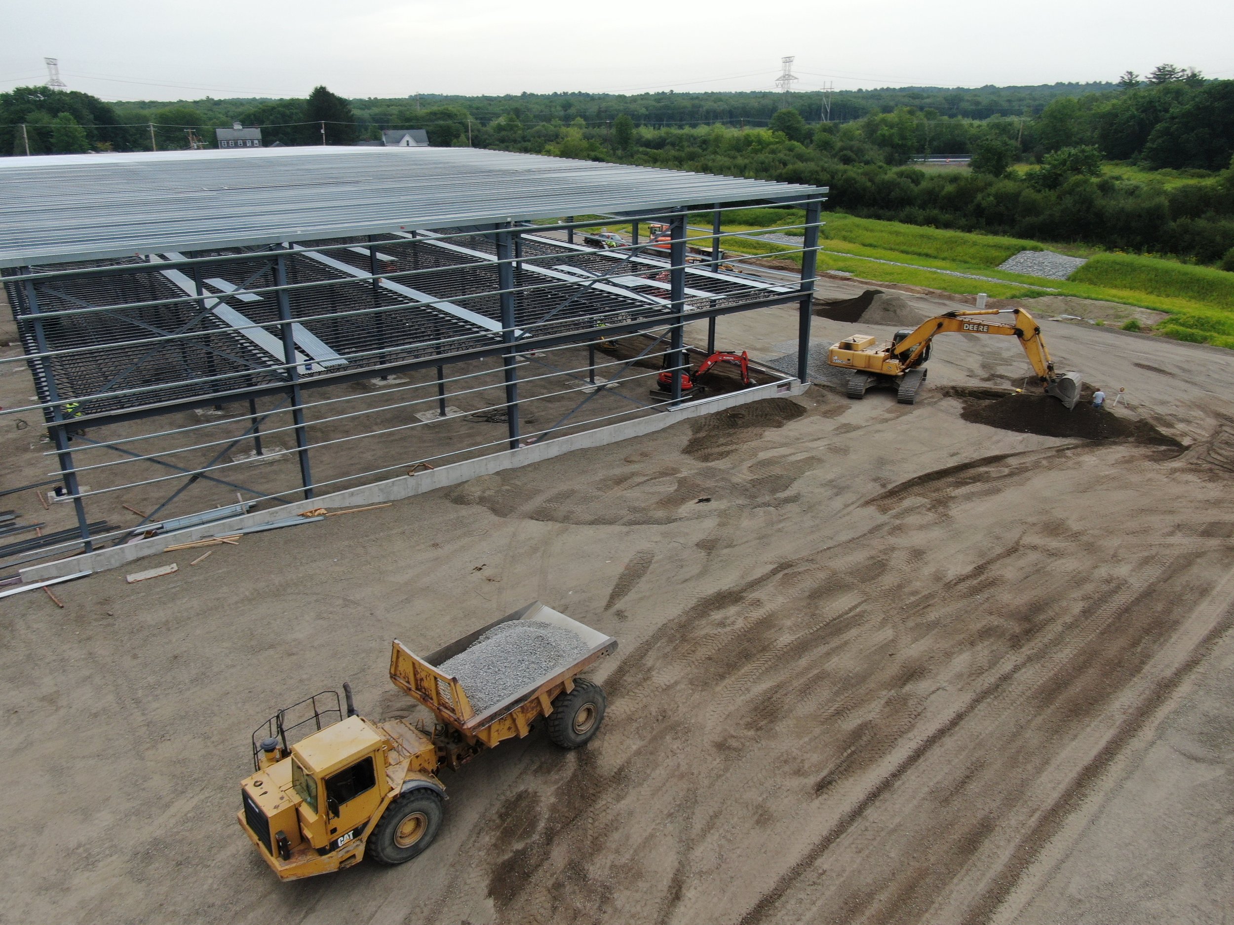 Construction site with a steel-framed building under construction, construction vehicles including a dump truck, an excavator, and a small roller. The area is dusty with piles of gravel.