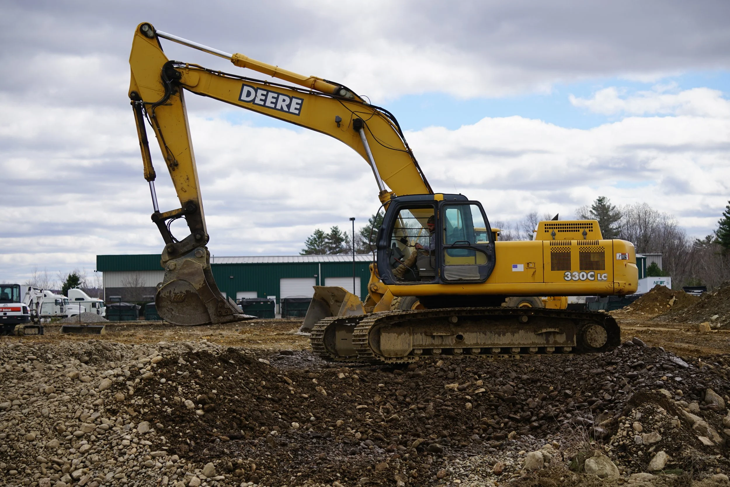 A yellow John Deere excavator digging into a construction site with piles of dirt and rocks, several trucks and a warehouse in the background under a cloudy sky.