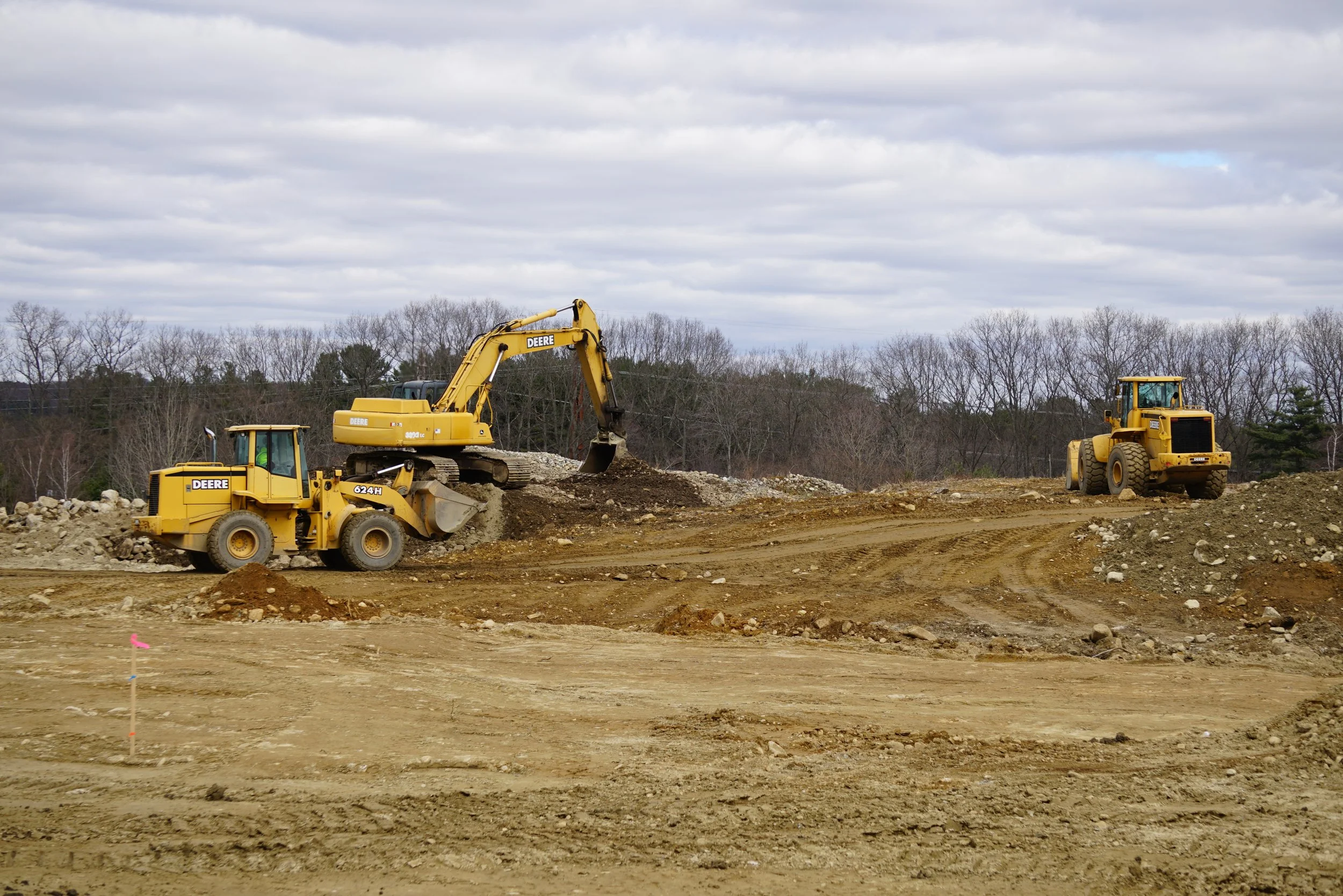 Construction site with two yellow heavy machinery vehicles, an excavator and a bulldozer, working on dirt and rocks under a cloudy sky.
