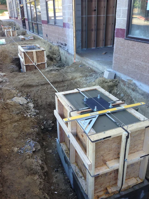 Construction site with concrete forms on ground, wood frames, and measurement tools, next to a building with large windows.