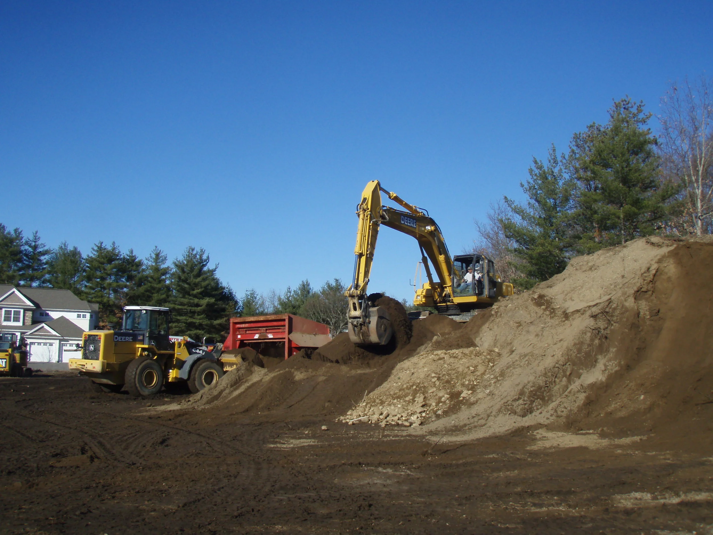 Construction site with a yellow excavator scooping dirt and a red trailer while multiple houses and trees are visible in the background under a blue sky.
