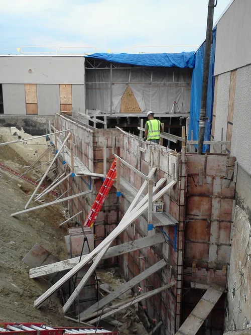 Construction worker in a neon safety vest and helmet working on a scaffolding wall supporting a building under construction, with wooden and steel supports and a partially covered site.