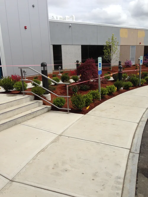 View of a sidewalk leading to the entrance of a commercial building, with landscaped flower beds, shrubs, and trees, and accessible parking signs.