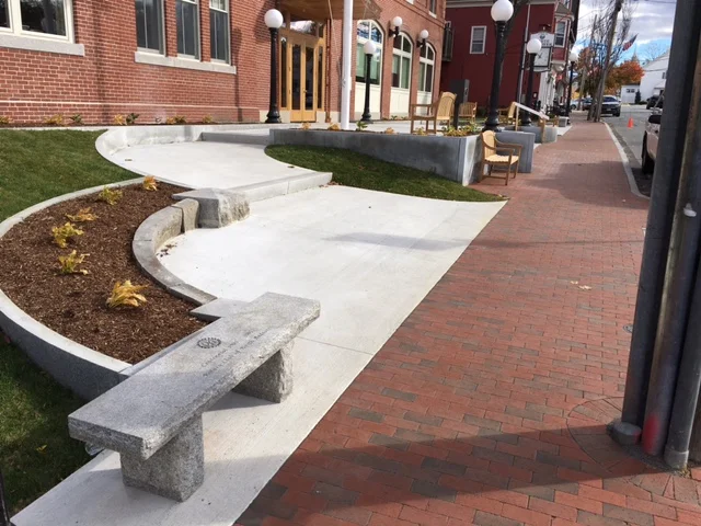 Sidewalk with a planter containing some small plants and fallen leaves, benches, and street lamps along a brick-paved sidewalk in an urban area.