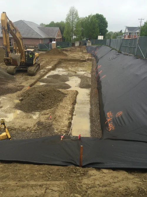 Construction site with excavated area, black waterproofing membrane on the ground, and a yellow excavator on the left. Residential houses and trees in the background.