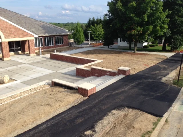 Sidewalk and driveway with patches of dirt and newly laid asphalt leading to a building with a brick exterior and large windows. Trees and residential houses are visible in the background.