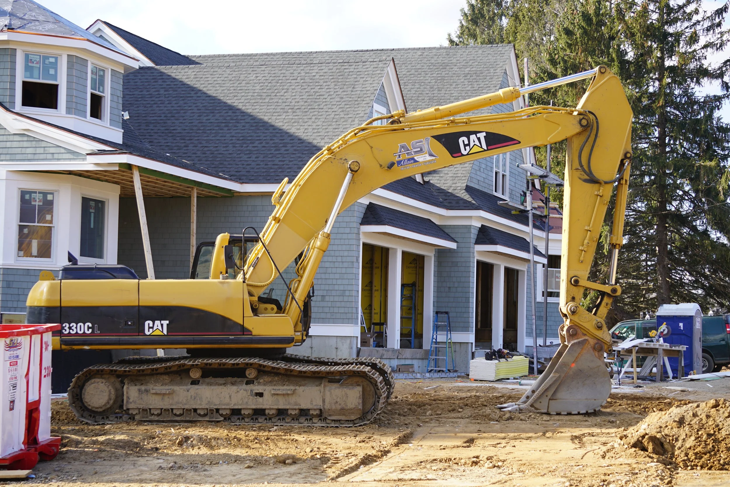 Construction site with a yellow CAT excavator working on a house renovation, with construction materials and scaffolding around the house.