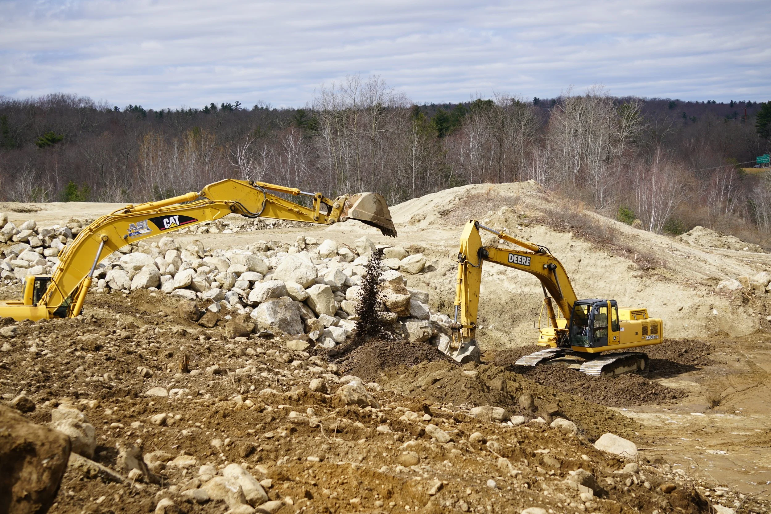 Construction site with two yellow excavators moving dirt and rocks, with a background of trees and a cloudy sky.