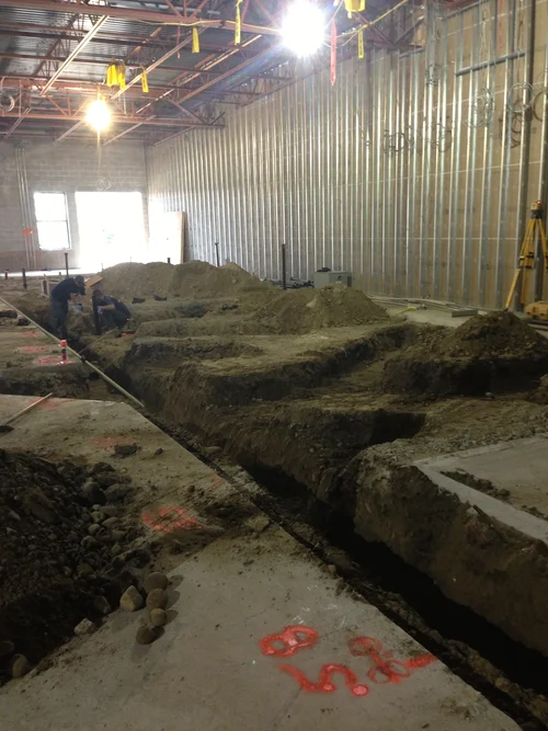 Construction workers working on underground utility installation inside a building with steel walls and ceiling, with piles of dirt and construction tools.