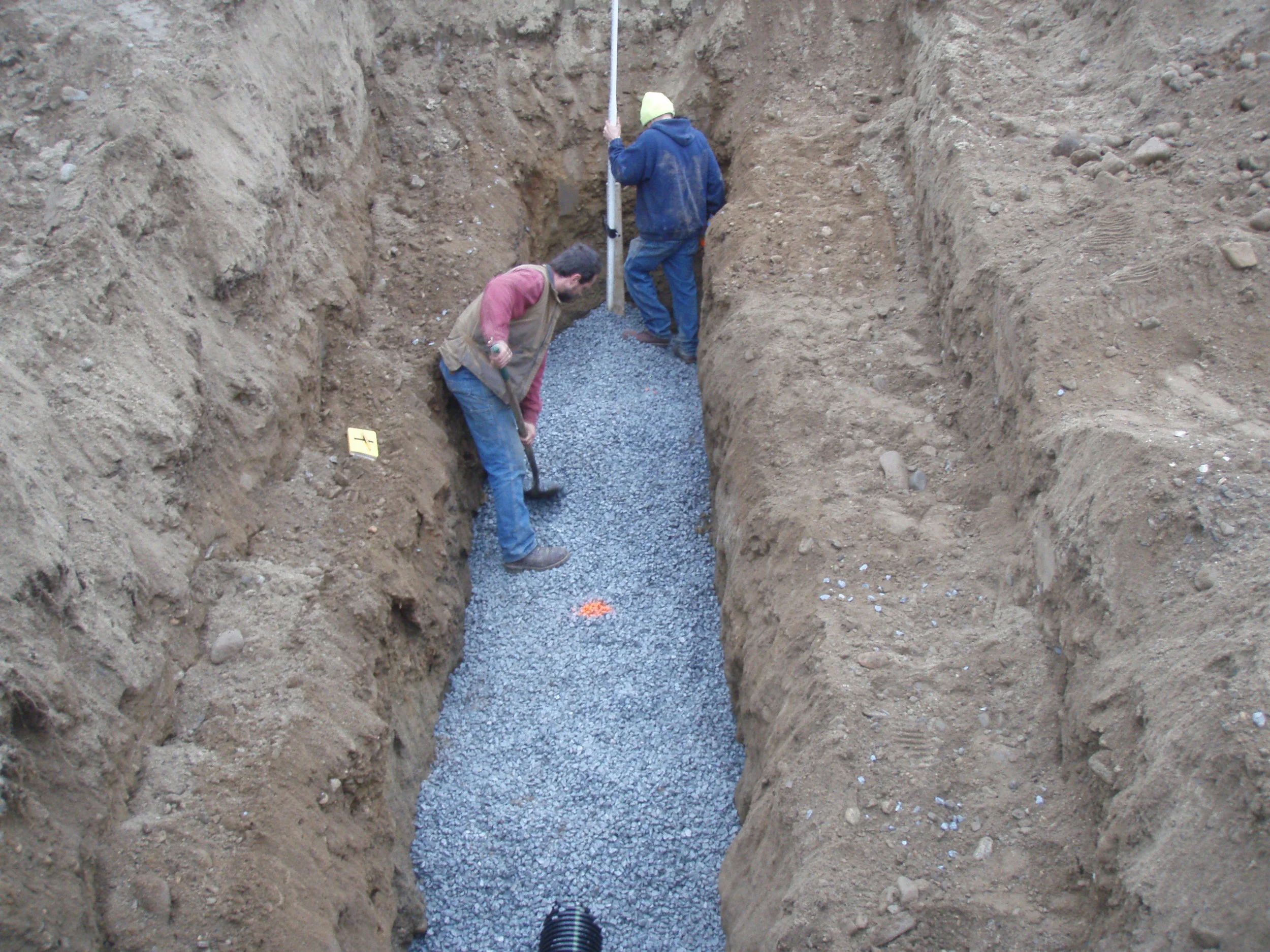 Two workers are working in a trench, spreading gravel evenly on the bottom of the trench. One worker is wearing a tan vest and the other a blue jacket and a yellow beanie. They are using shovels and a leveling rod.
