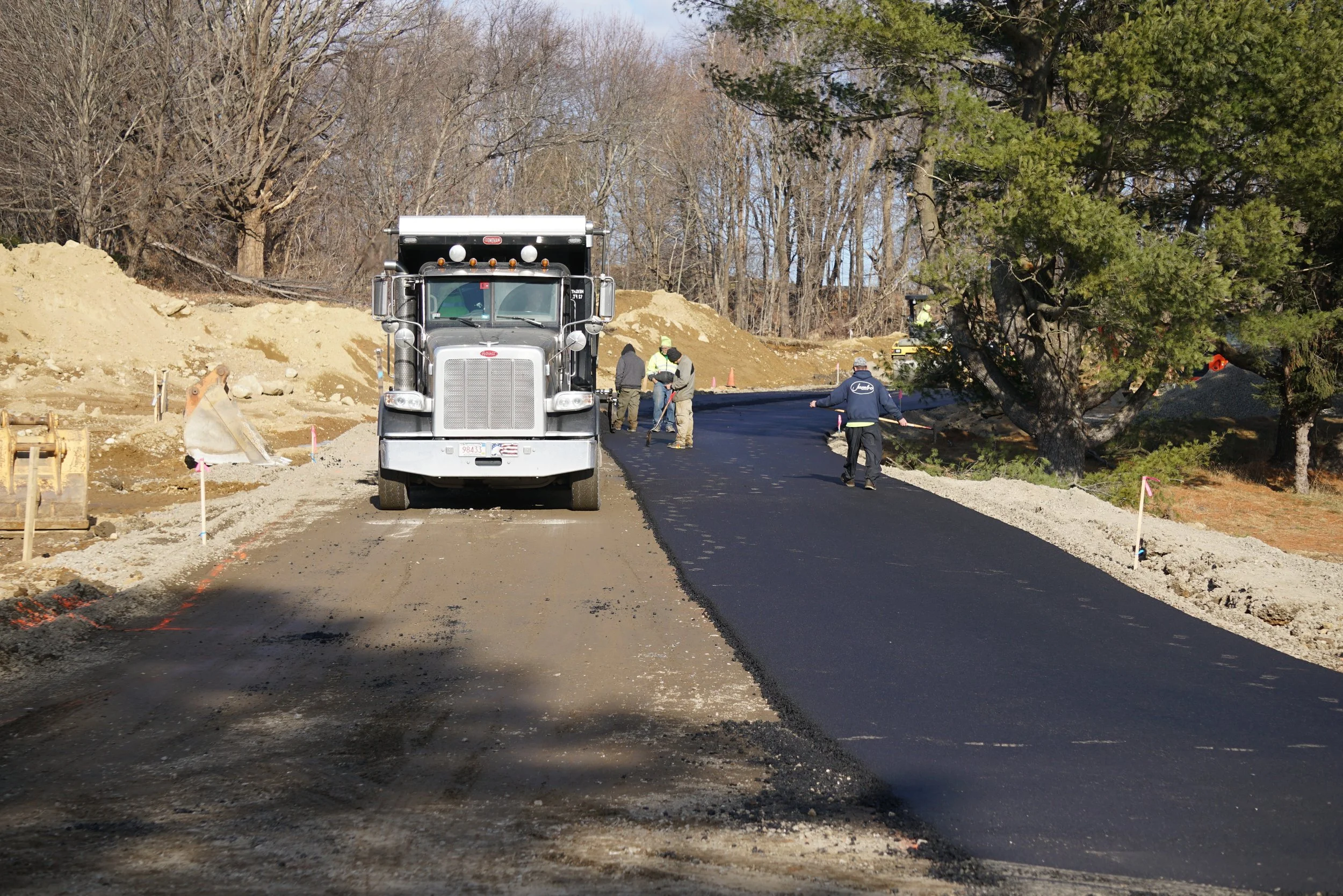 Workers paving a road with asphalt, with construction equipment and large trees in the background.