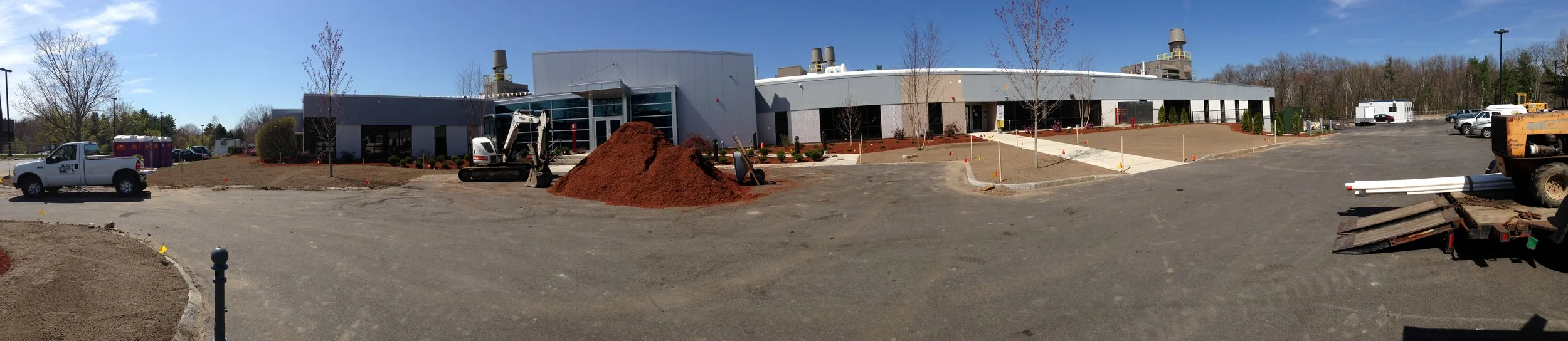 Construction site in front of a commercial building with trees and vehicles in the background, and construction materials and equipment in the foreground.