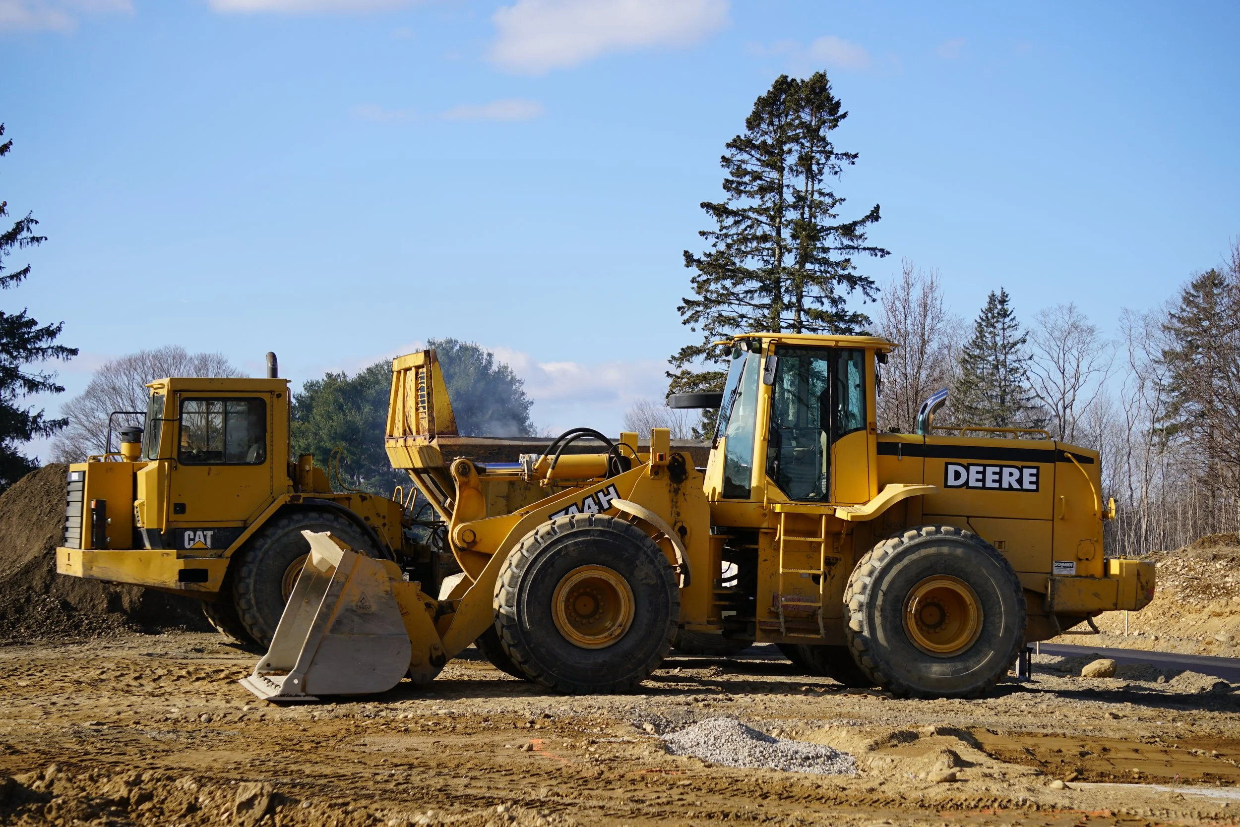 A yellow bulldozer on construction site with dirt and a large tree in the background, under a partly cloudy sky.