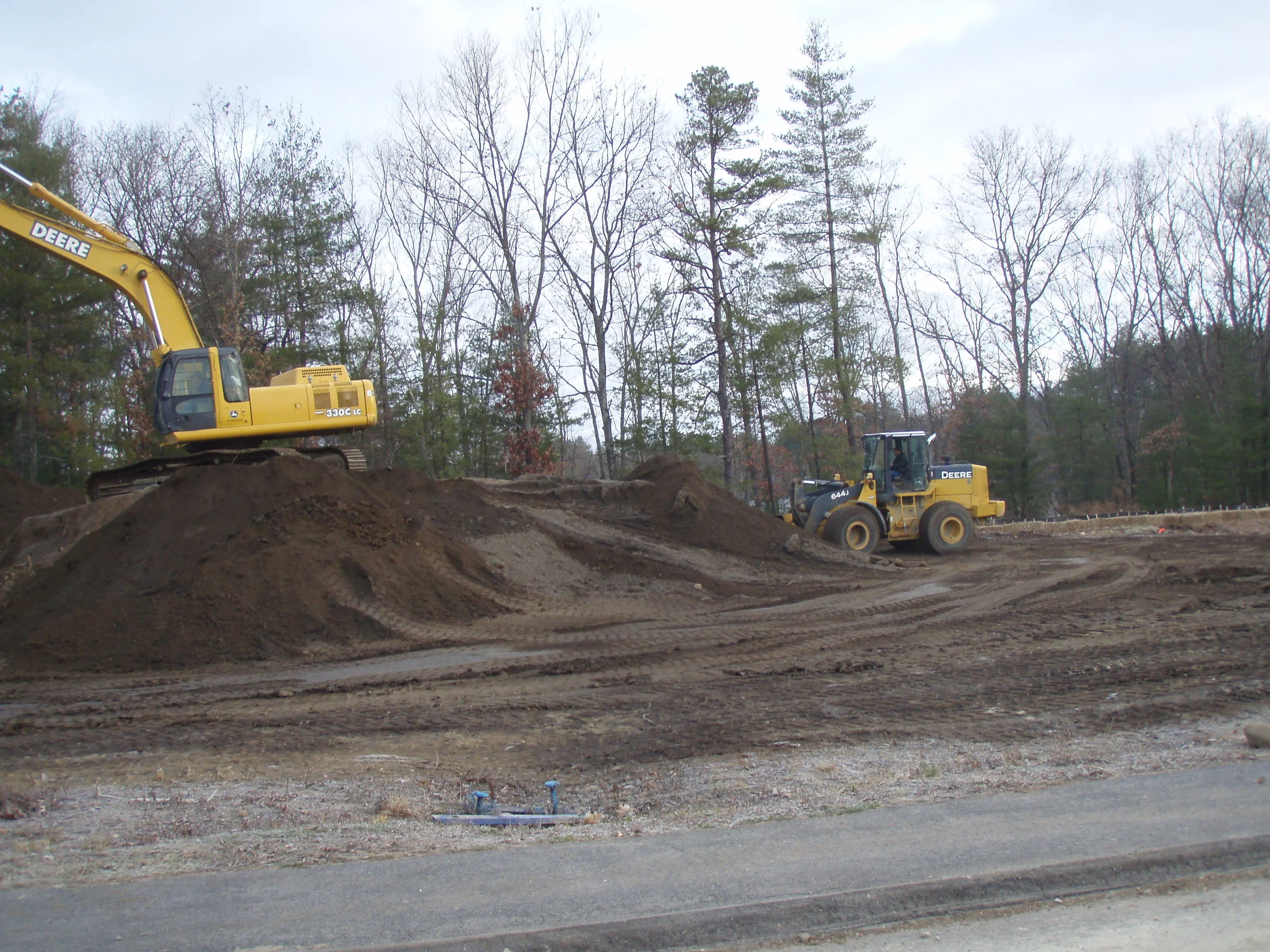Construction site with a yellow excavator digging in dirt and a yellow compact loader machine, with a background of trees and a cloudy sky.