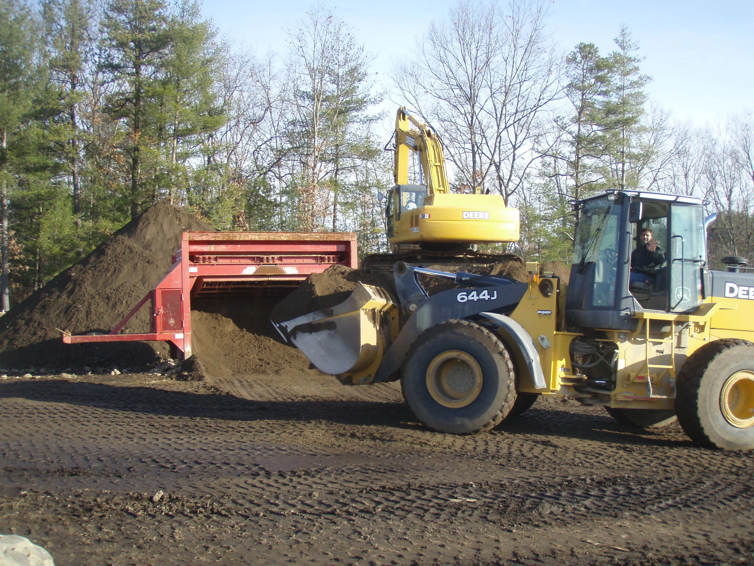 Construction site with a yellow excavator moving dirt into a red truck, surrounded by trees.