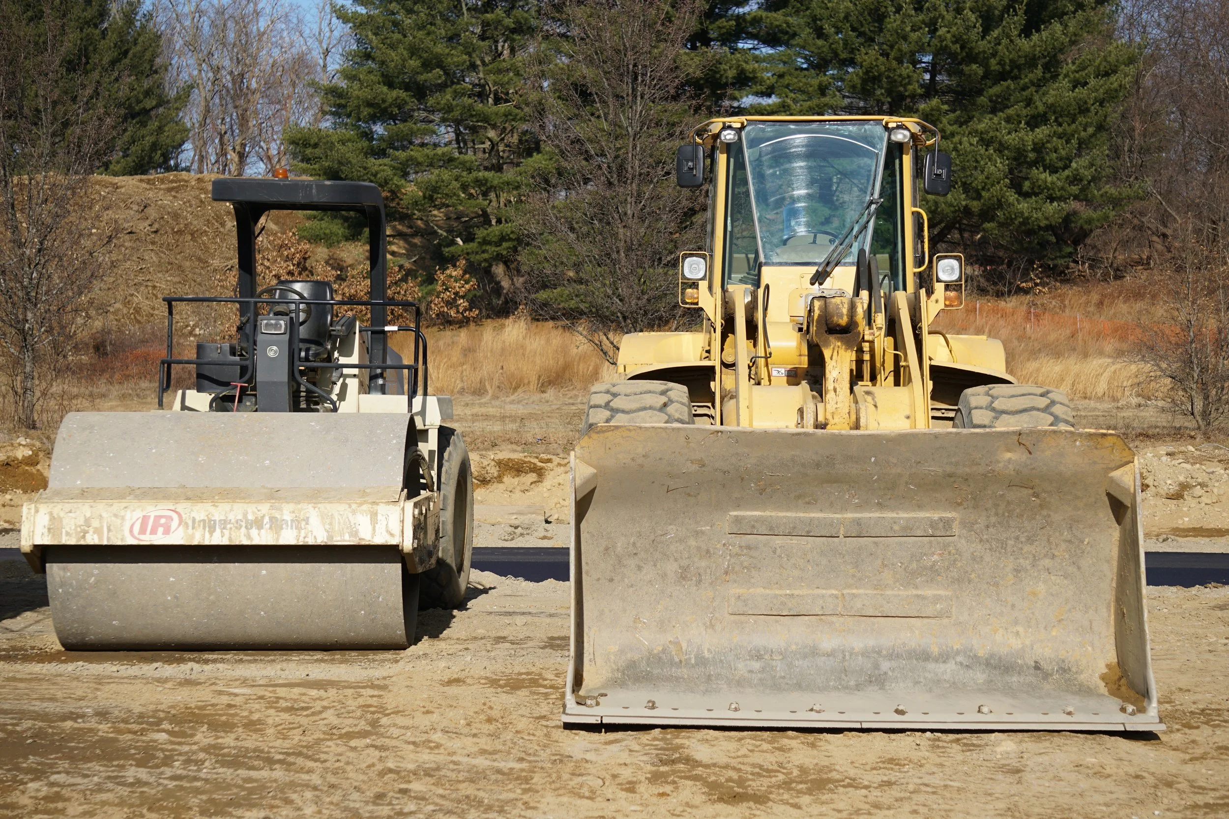 Construction site with two large rollers and a yellow bulldozer in a rural area with trees and grass.