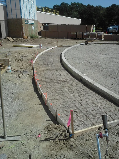 Construction site with a curved pathway being built, with rebar and formwork in place, and workers and materials in the background.